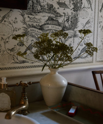 A white Cirrus Bud Vase with green foliage sits on a bathroom counter beside a sink, soap dispenser, and bird-themed vintage wallpaper.