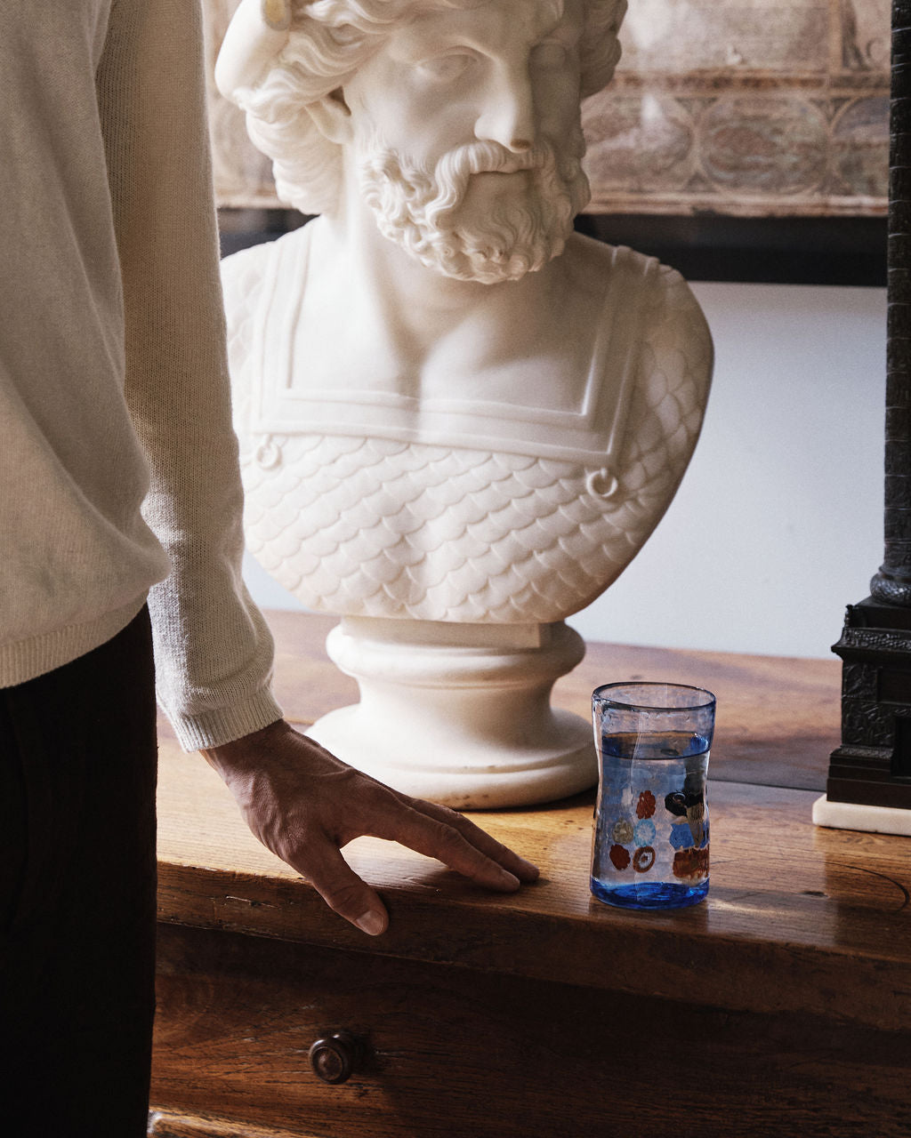 A person stands beside a wooden table with a Muranesi Glass, Set of 2 in blue and a white marble bust of a bearded man placed on it.
