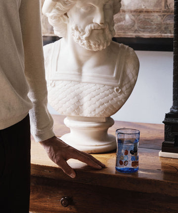 A person stands beside a wooden table with a Muranesi Glass, Set of 2 in blue and a white marble bust of a bearded man placed on it.