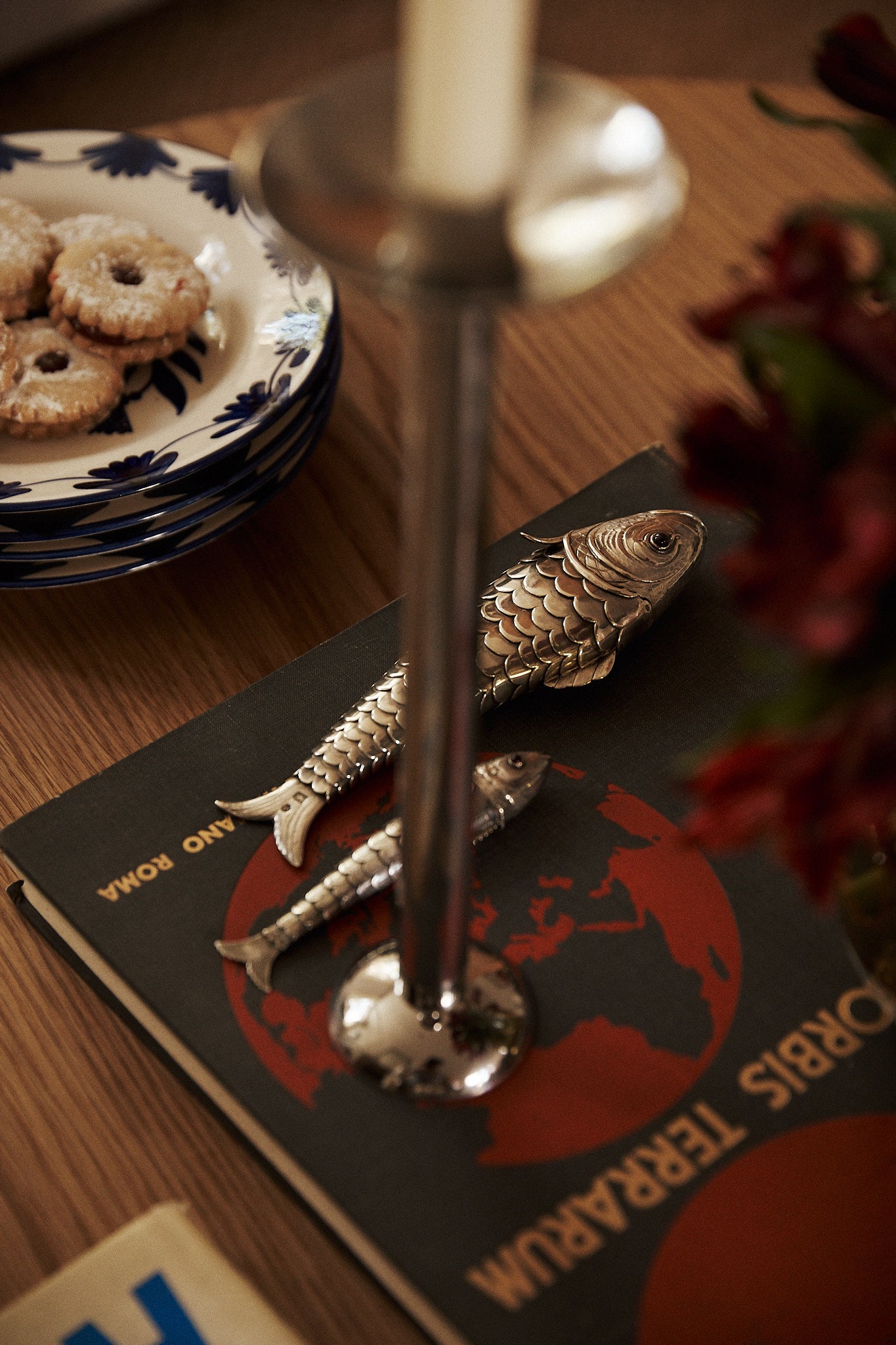 A candlestick and two vintage silver fish figurines rest on a book with a red map cover; the Vintage Silver Articulating Fish serves as a lucky talisman nearby, while cookies on a floral plate and blurred flowers are also visible.