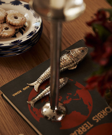 A candlestick and two vintage silver fish figurines rest on a book with a red map cover; the Vintage Silver Articulating Fish serves as a lucky talisman nearby, while cookies on a floral plate and blurred flowers are also visible.