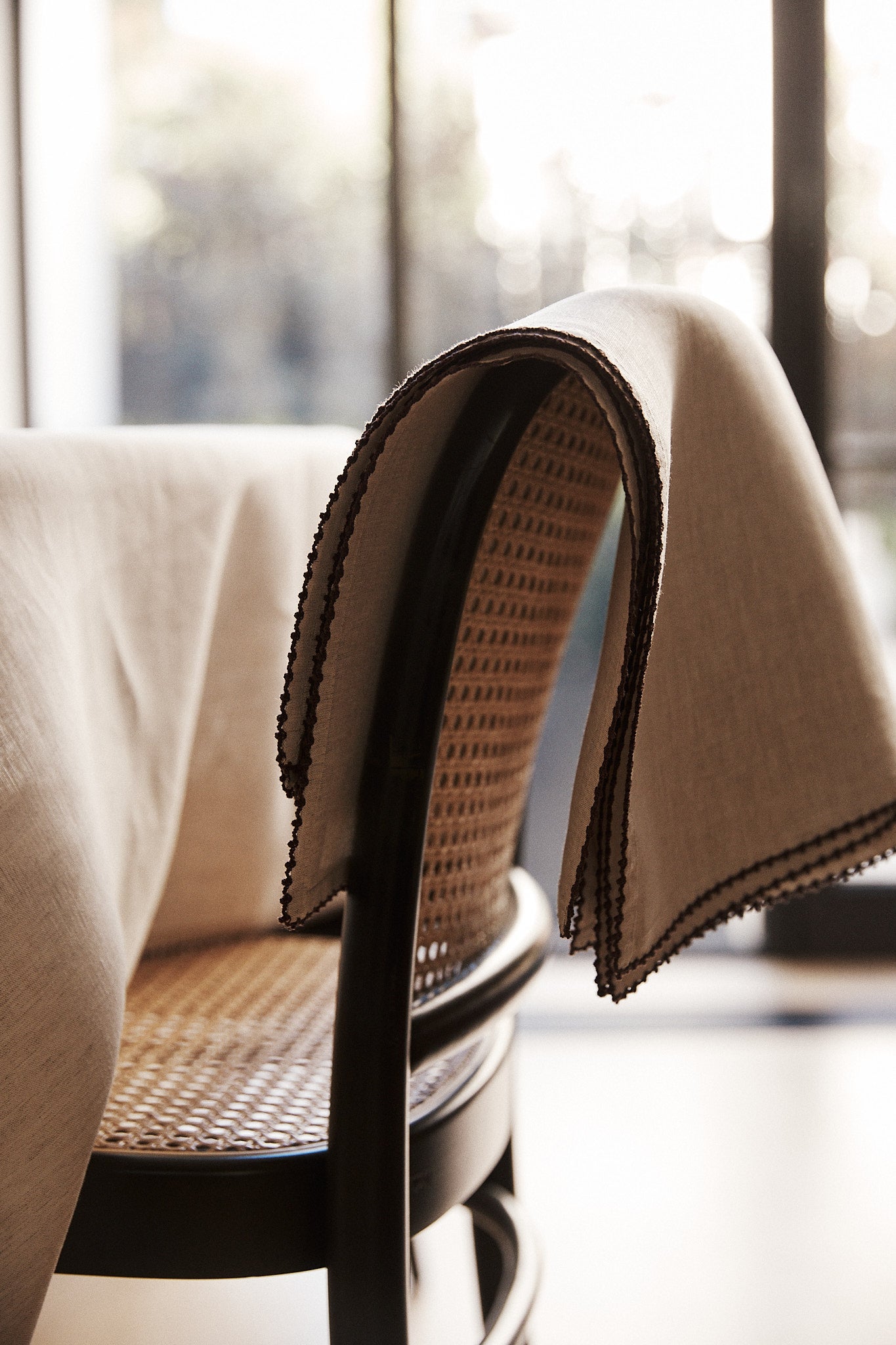 A set of Nico Placemats in beige, featuring hand-stitched edges, is draped over the back of a woven cane chair beside a table, with large windows in the background.