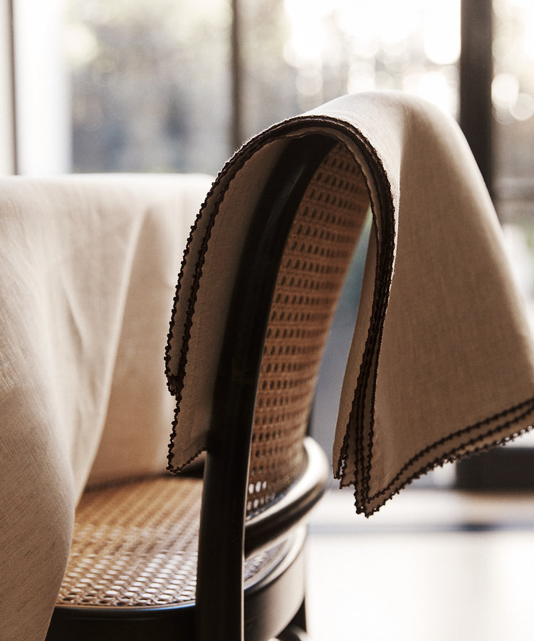 A set of Nico Placemats in beige, featuring hand-stitched edges, is draped over the back of a woven cane chair beside a table, with large windows in the background.