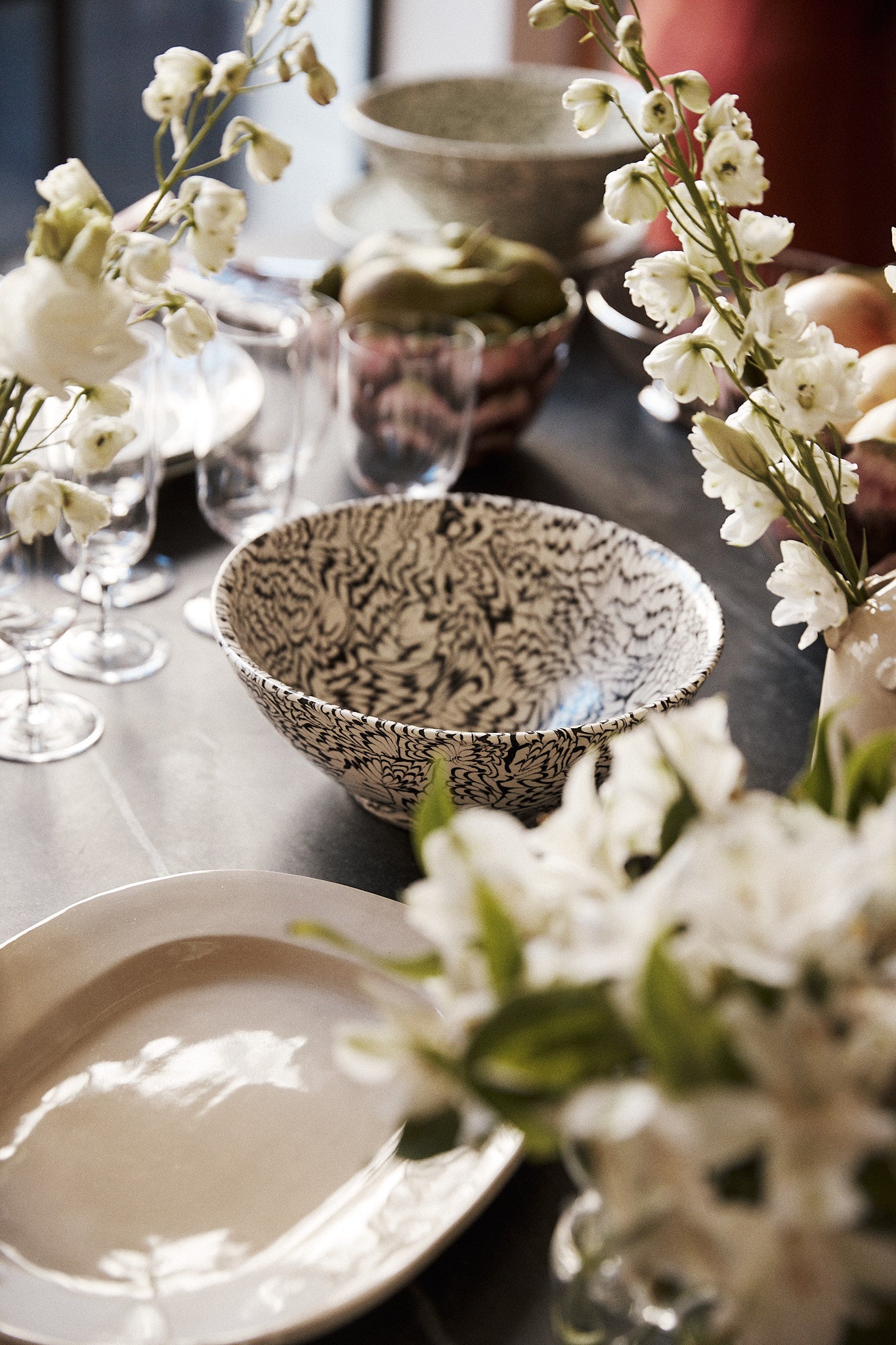 A close-up of a dining table set with plates, glasses, the intricately patterned Celine Salad Bowl, and vases of white flowers.