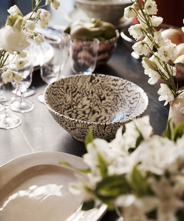 A close-up of a dining table set with plates, glasses, the intricately patterned Celine Salad Bowl, and vases of white flowers.