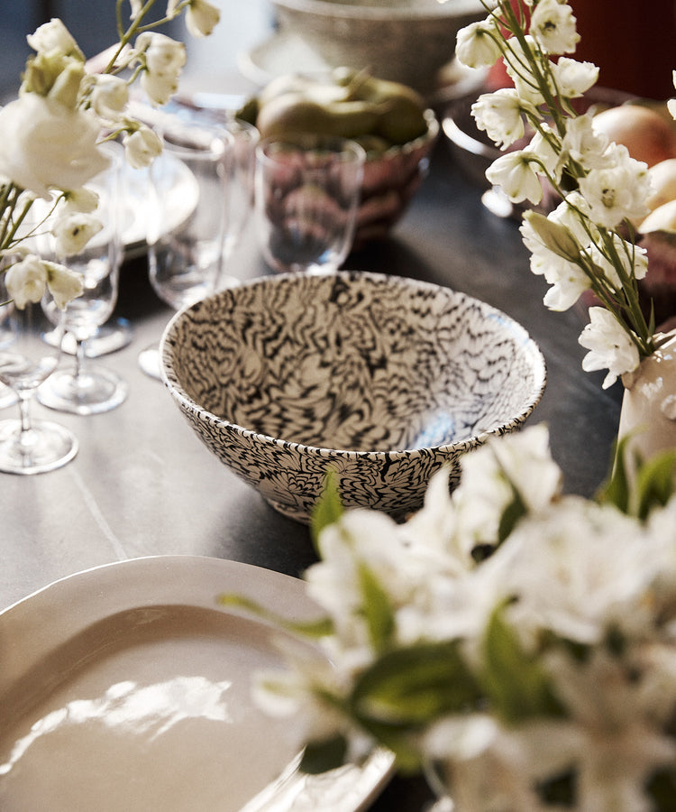 A close-up of a dining table set with plates, glasses, the intricately patterned Celine Salad Bowl, and vases of white flowers.