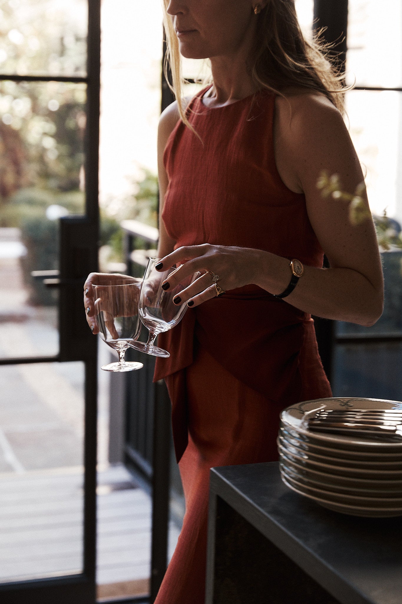 A woman in a sleeveless rust-colored dress holds two Venezia Wine Glasses from the Set of 6 near a stack of plates in a sunlit room with glass doors.