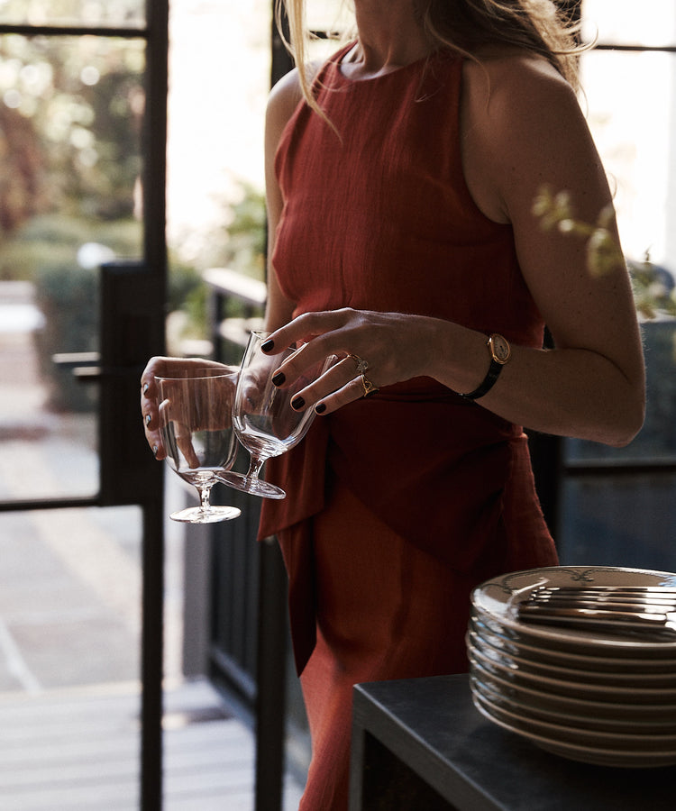 A woman in a sleeveless rust-colored dress holds two Venezia Wine Glasses from the Set of 6 near a stack of plates in a sunlit room with glass doors.