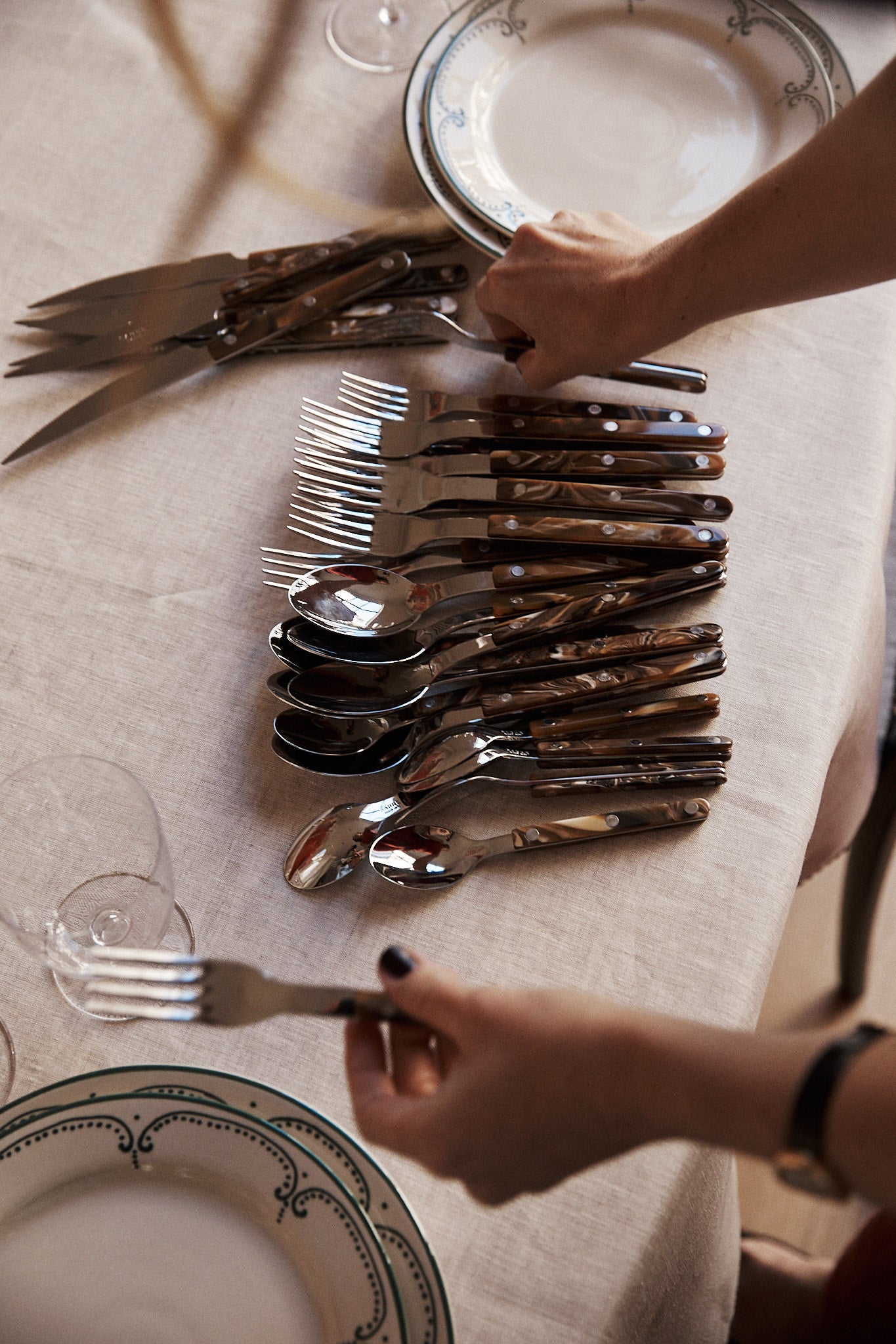 A person sets a table with the Bistrot 5 Piece Cutlery Set and plates on a white tablecloth; the cutlery, featuring buffalo-style handles, is arranged in rows, and one fork from the set is being held.