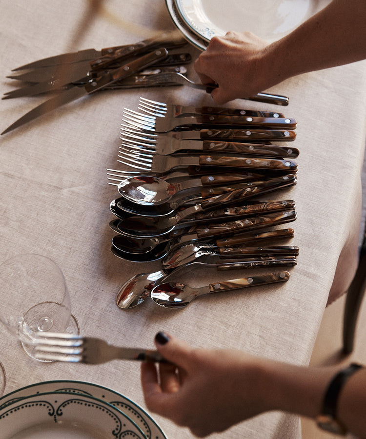 A person sets a table with the Bistrot 5 Piece Cutlery Set and plates on a white tablecloth; the cutlery, featuring buffalo-style handles, is arranged in rows, and one fork from the set is being held.