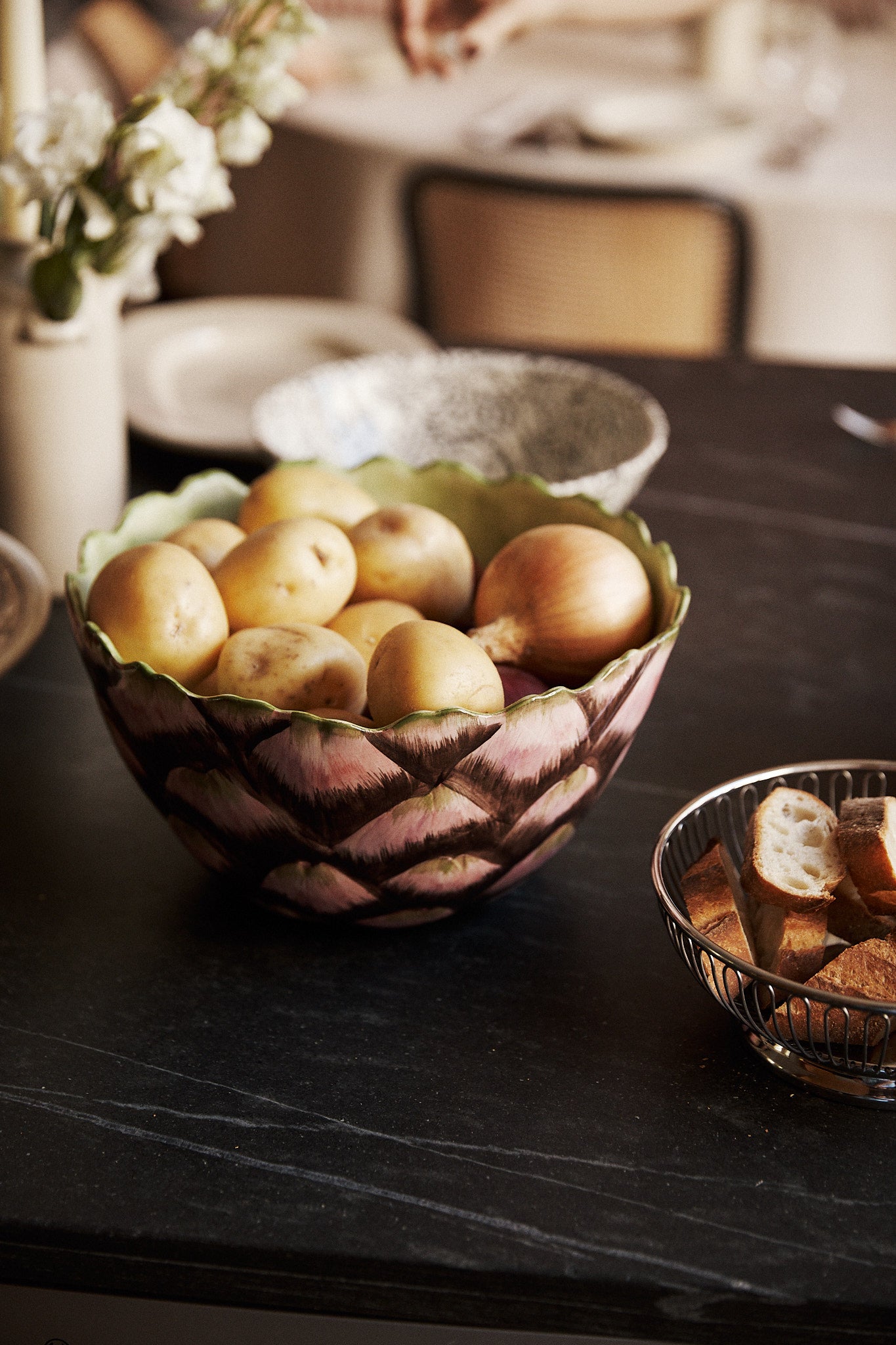 An Artichoke Serving Bowl—crafted from hand-finished ceramic—holds a pile of potatoes atop a dark countertop, its artisanal charm evocative of traditional Italian clayware, while a metal basket nearby contains sliced bread.