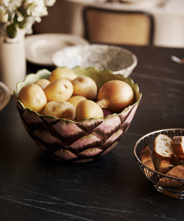 An Artichoke Serving Bowl—crafted from hand-finished ceramic—holds a pile of potatoes atop a dark countertop, its artisanal charm evocative of traditional Italian clayware, while a metal basket nearby contains sliced bread.