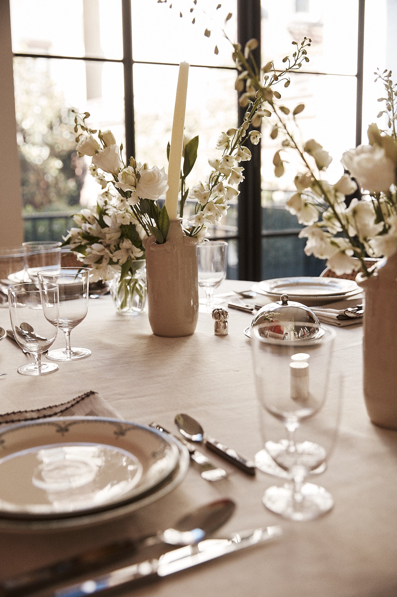 A dining table set with plates, silverware, glassware, and a Mini Candlehat Vase holding white flowers, plus a candle holder, all elegantly arranged near large windows.