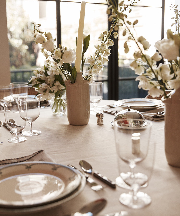 A dining table set with plates, silverware, glassware, and a Mini Candlehat Vase holding white flowers, plus a candle holder, all elegantly arranged near large windows.
