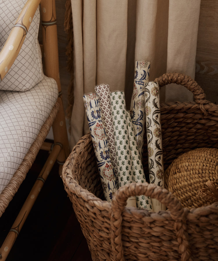 A woven basket holds several rolled sheets of Rampant Lions Gift Wrap and a round, woven object. A cushioned chair and curtain are partially visible beside the basket.