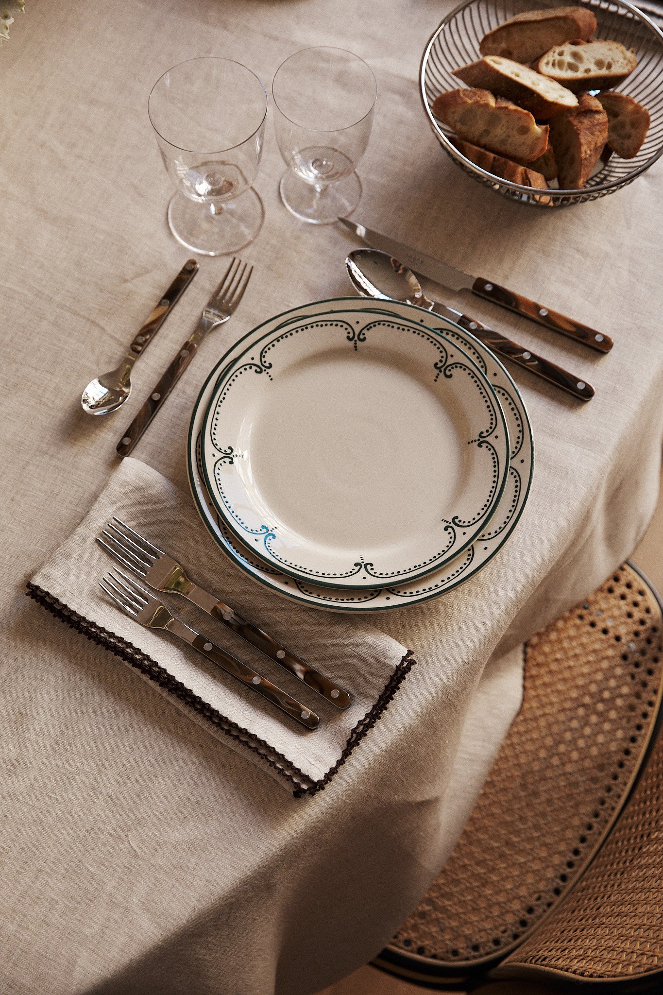 A table set with a decorative plate, cutlery on Nico Napkin, Set of 6, two wine glasses, and a basket of sliced bread arranged on a beige tablecloth.