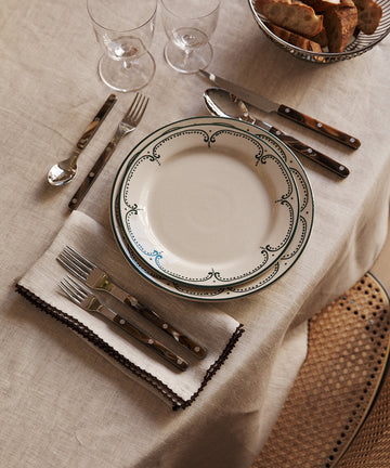 A table set with a decorative plate, cutlery on Nico Napkin, Set of 6, two wine glasses, and a basket of sliced bread arranged on a beige tablecloth.