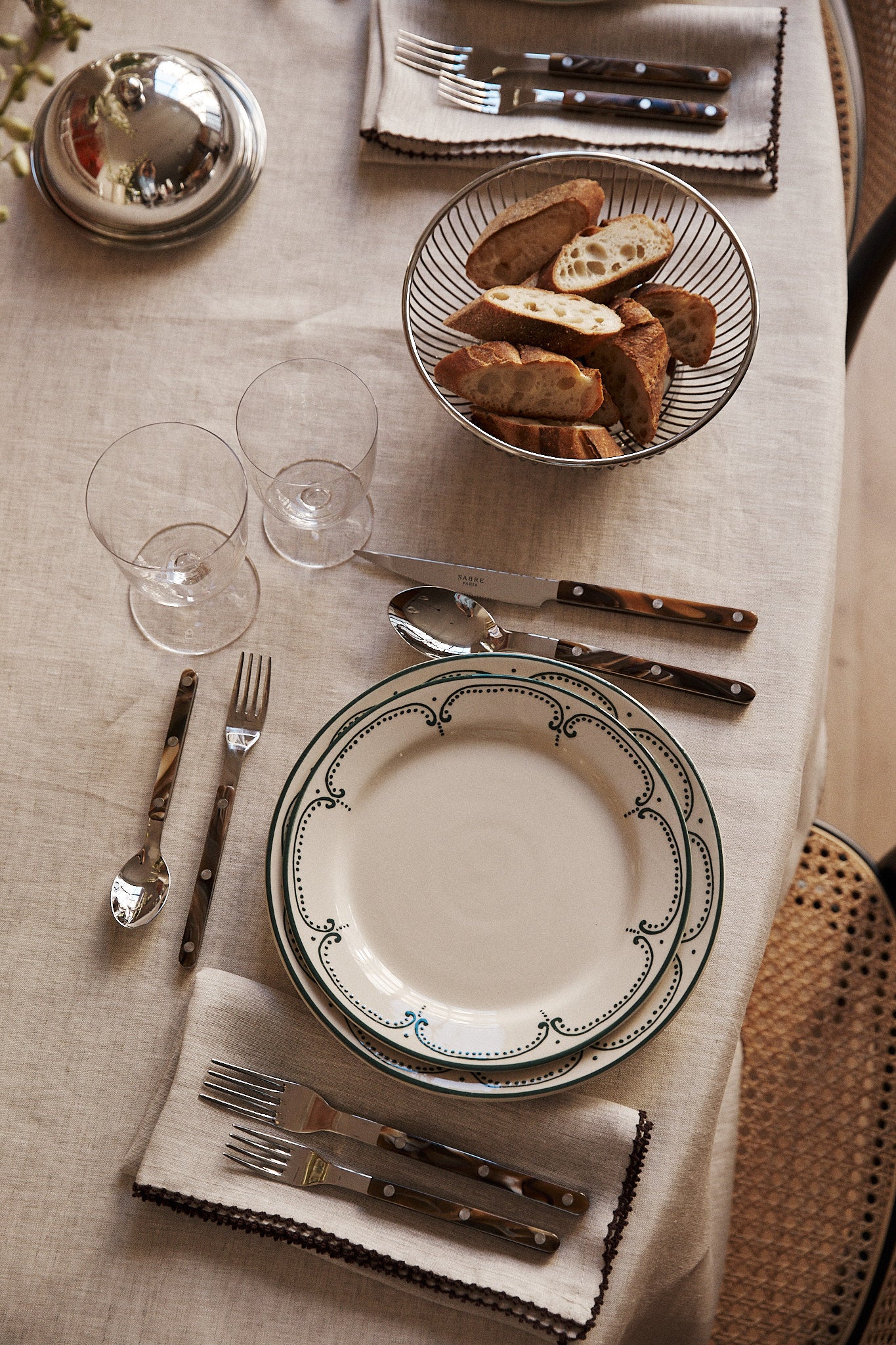 A table set with an Arco Dinner Plate, cutlery, two empty glasses, napkins, and a bowl of sliced bread on a beige tablecloth.