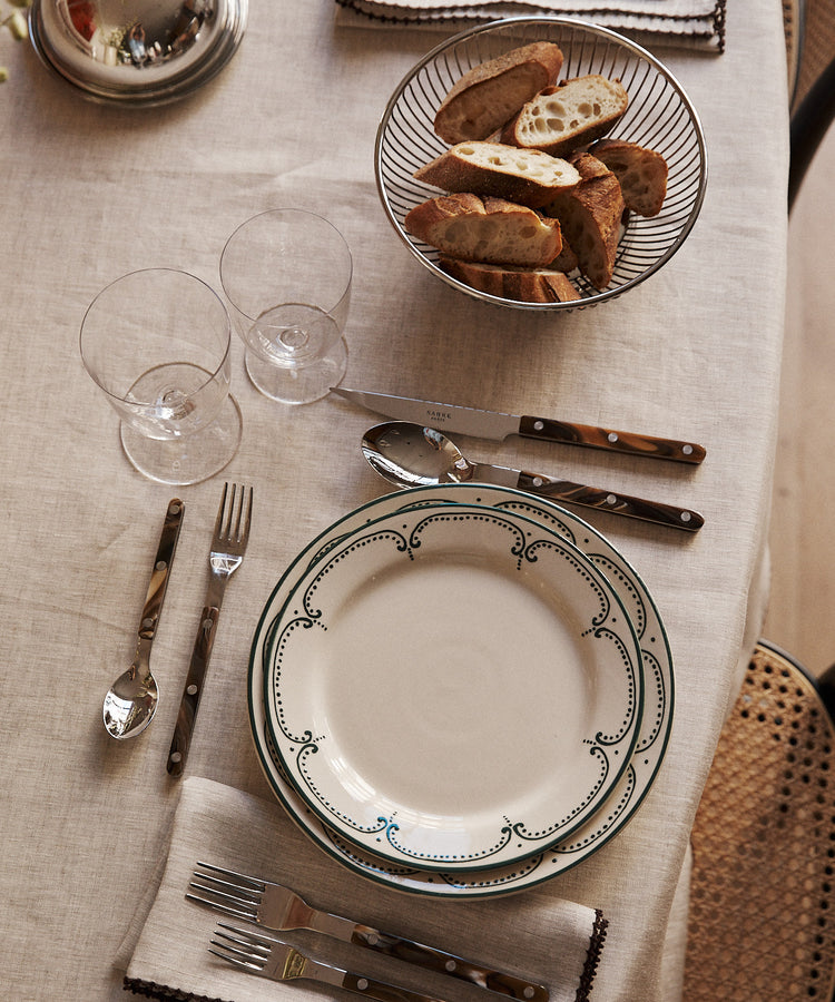 A table set with an Arco Dinner Plate, cutlery, two empty glasses, napkins, and a bowl of sliced bread on a beige tablecloth.