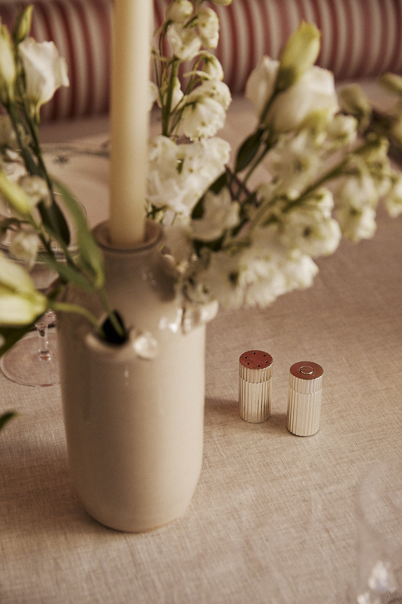 The Sophia Pepper Mill and Salt Shaker, crafted with refined Italian artistry, sit on a beige tablecloth beside a ceramic vase with white flowers and a tall candle.