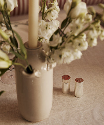The Sophia Pepper Mill and Salt Shaker, crafted with refined Italian artistry, sit on a beige tablecloth beside a ceramic vase with white flowers and a tall candle.