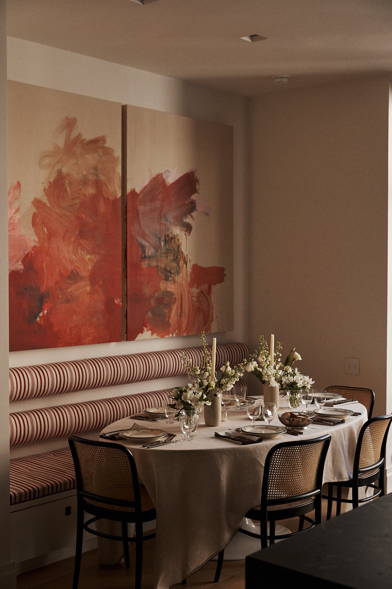 The dining table is dressed with the Nico Tablecloth, set for a meal and surrounded by chairs and a striped banquette, with floral centerpieces and abstract art on the wall.