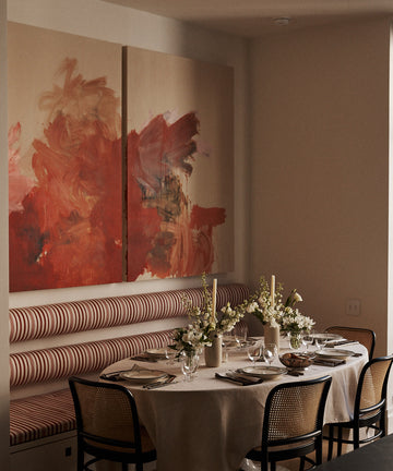 The dining table is dressed with the Nico Tablecloth, set for a meal and surrounded by chairs and a striped banquette, with floral centerpieces and abstract art on the wall.