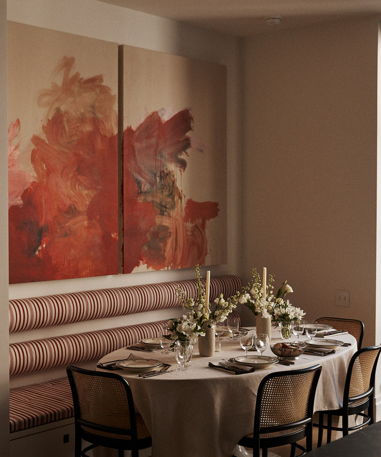 The dining table is dressed with the Nico Tablecloth, set for a meal and surrounded by chairs and a striped banquette, with floral centerpieces and abstract art on the wall.