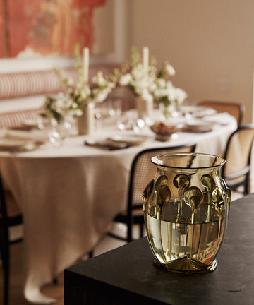 A textured, translucent gold glass vase with a floral pattern sits on a dark table in a softly blurred dining room setting.