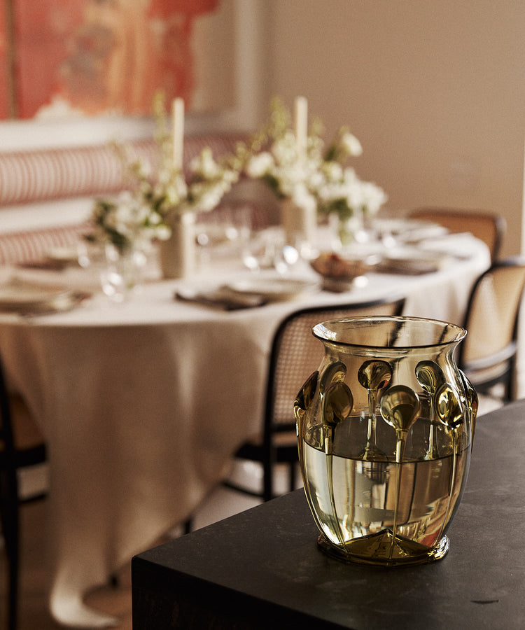 A textured, translucent gold glass vase with a floral pattern sits on a dark table in a softly blurred dining room setting.