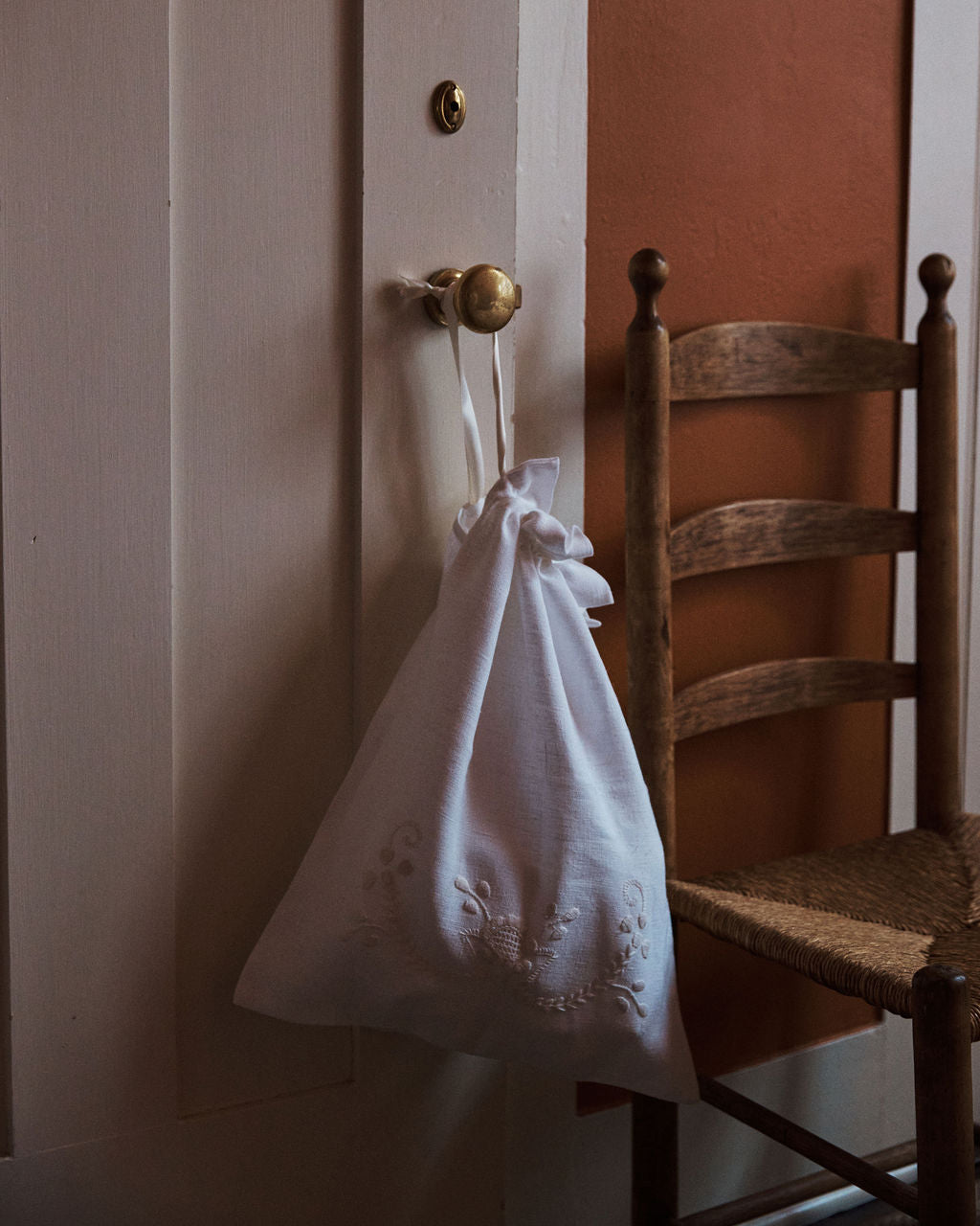 The Linen Drawstring Bag, handmade in Portugal and inspired by Viana do Castelo embroidery, hangs from a brass door knob beside a wooden chair with a woven seat.