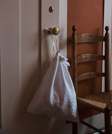 The Linen Drawstring Bag, handmade in Portugal and inspired by Viana do Castelo embroidery, hangs from a brass door knob beside a wooden chair with a woven seat.