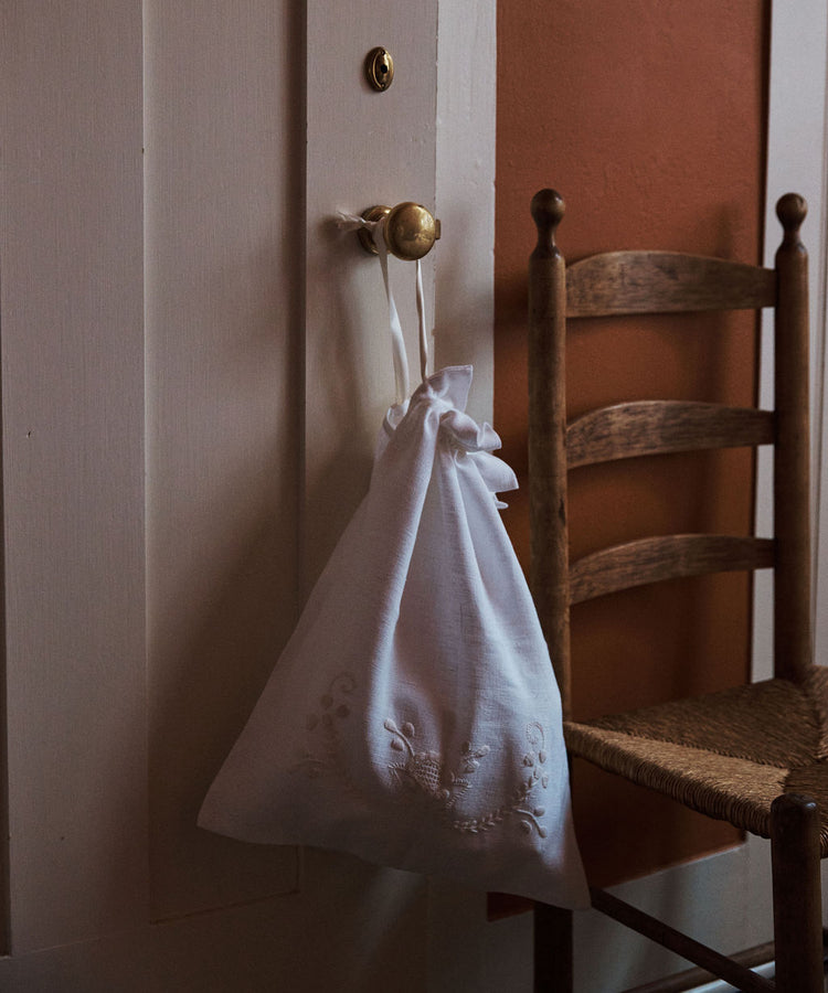 The Linen Drawstring Bag, handmade in Portugal and inspired by Viana do Castelo embroidery, hangs from a brass door knob beside a wooden chair with a woven seat.