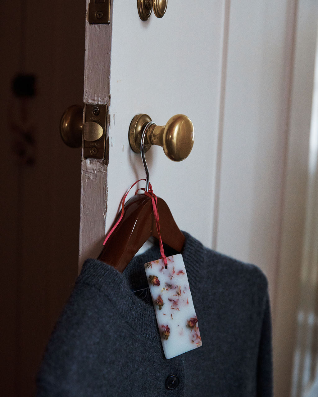 A gray sweater on a wooden hanger hangs from a brass doorknob, with a Rosa Novella Scented Wax Tablet—attached by a red ribbon—adding a subtle Tuscan fragrance to the air.
