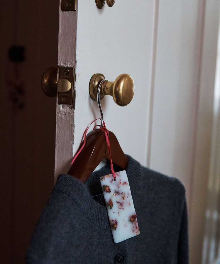 A gray sweater on a wooden hanger hangs from a brass doorknob, with a Rosa Novella Scented Wax Tablet—attached by a red ribbon—adding a subtle Tuscan fragrance to the air.