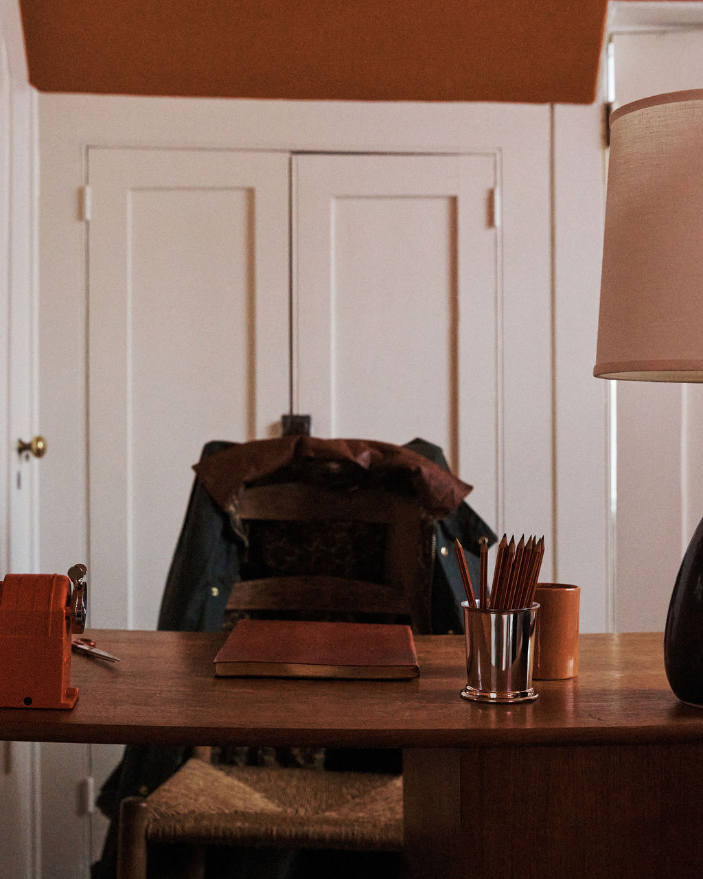 The Red Desk Bundle features a desk with accessories, a red leather journal, Viarco pencils in a holder, lamp, and sharpener, set before a chair with a jacket; white double doors appear in the background.