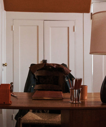 The Red Desk Bundle features a desk with accessories, a red leather journal, Viarco pencils in a holder, lamp, and sharpener, set before a chair with a jacket; white double doors appear in the background.