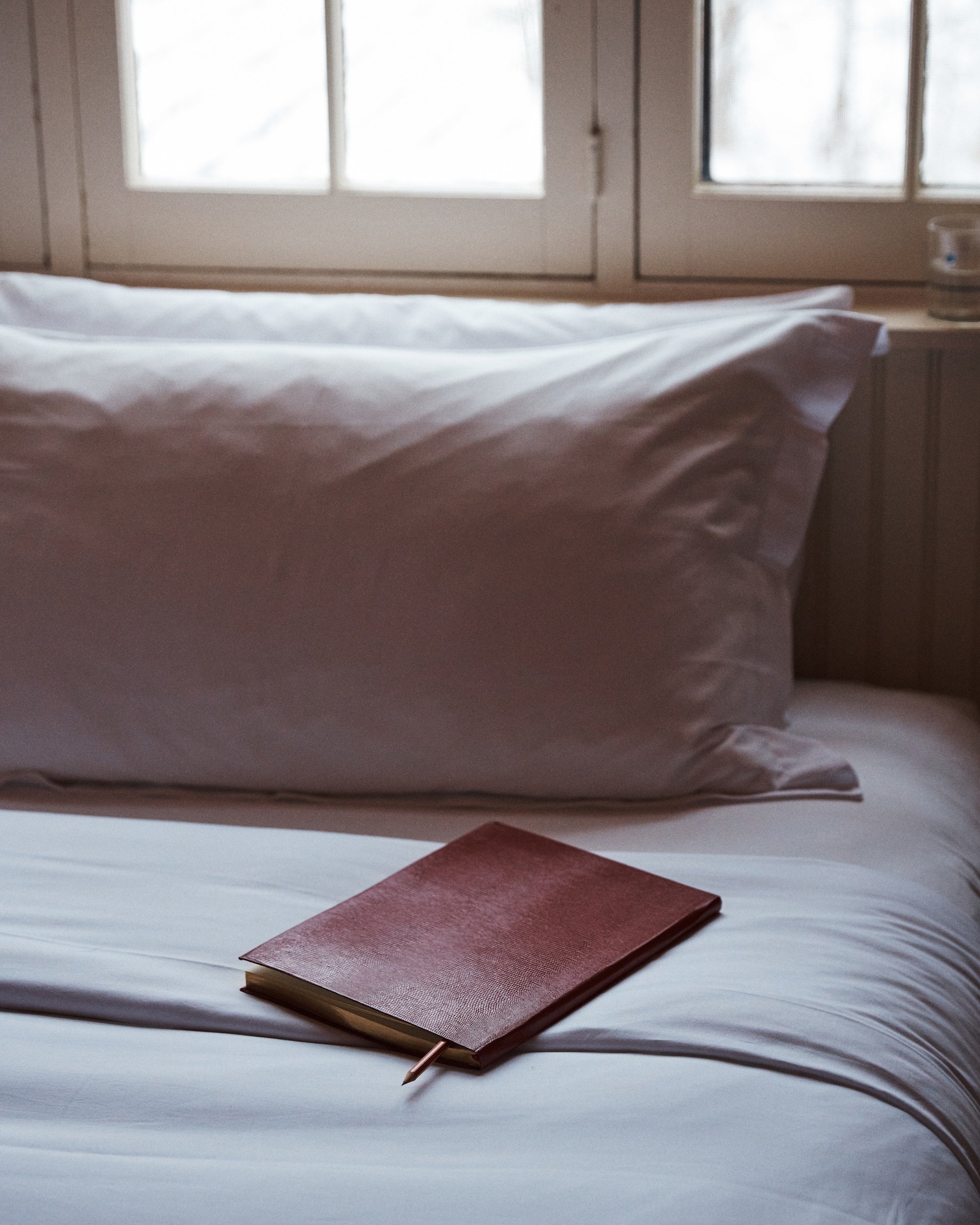 A closed, handcrafted Red Notebook with a lizard-embossed cover and pencil lies on a neatly made bed with white sheets and pillows, next to a window with frosted glass.