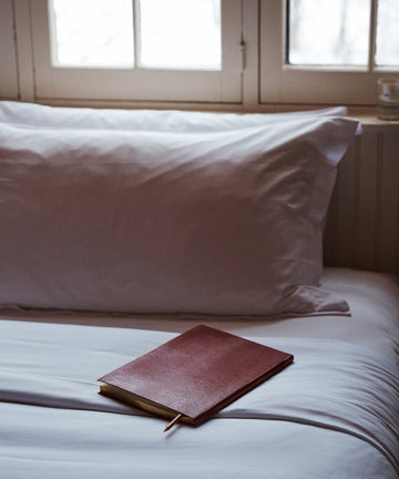A closed, handcrafted Red Notebook with a lizard-embossed cover and pencil lies on a neatly made bed with white sheets and pillows, next to a window with frosted glass.