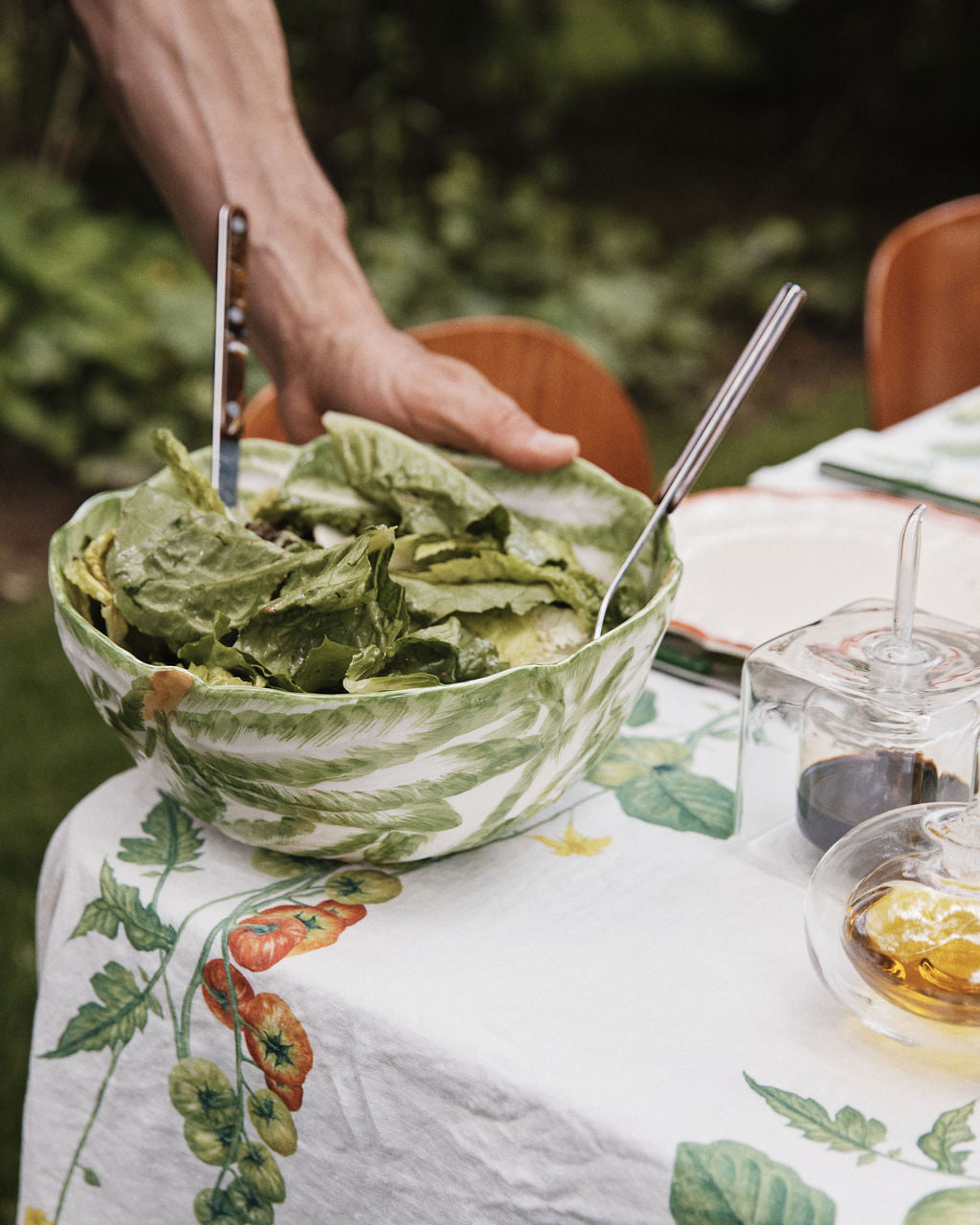 A hand holds a green salad in the Radicchio Serving Bowl, with serving utensils, atop a table draped in a vegetable-printed tablecloth.