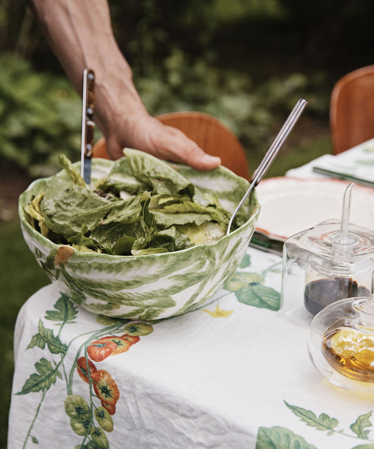 A hand holds a green salad in the Radicchio Serving Bowl, with serving utensils, atop a table draped in a vegetable-printed tablecloth.