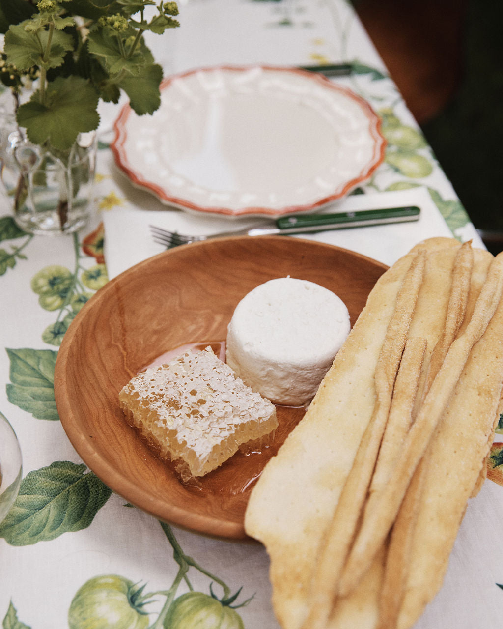 An August Wood Serving Bowl adds a touch of elegance to the inviting spread on the table, where a wooden plate presents a round of cheese, a piece of honeycomb, and two long bread crisps atop a charming tomato-patterned tablecloth.