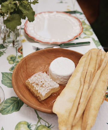 An August Wood Serving Bowl adds a touch of elegance to the inviting spread on the table, where a wooden plate presents a round of cheese, a piece of honeycomb, and two long bread crisps atop a charming tomato-patterned tablecloth.