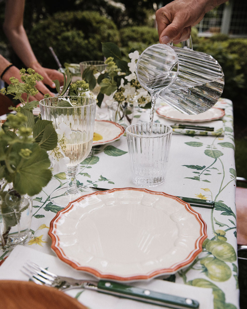 A hand pours water from the durable Fresco Pitcher into a tumbler on a table set with plates, utensils, wine glasses, and green floral decorations.