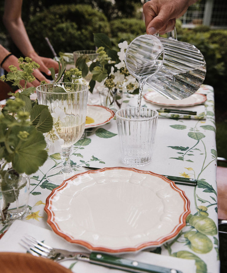A hand pours water from the durable Fresco Pitcher into a tumbler on a table set with plates, utensils, wine glasses, and green floral decorations.