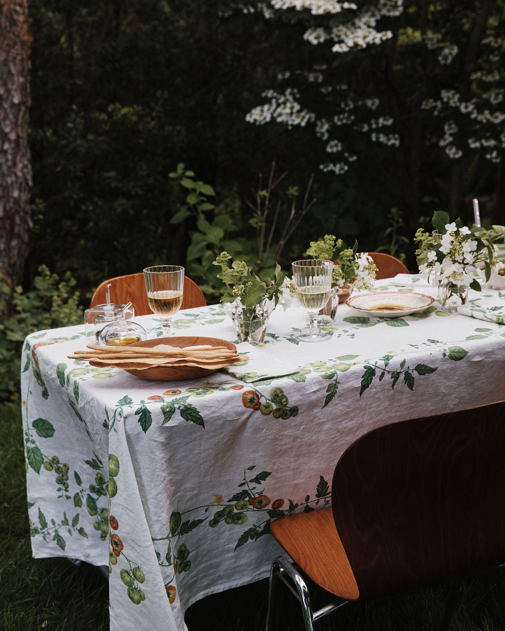 A table set outdoors with a Tomate Verte Linen Tablecloth, glassware, plates, and vases of fresh flowers, surrounded by wooden chairs and lush greenery.