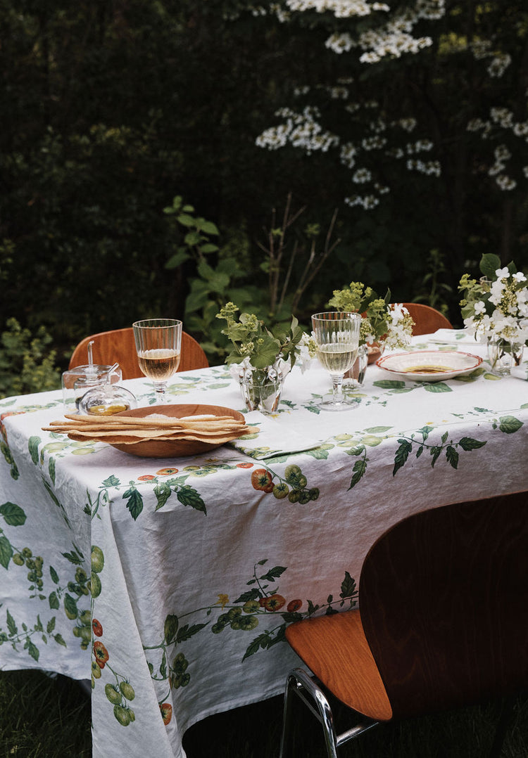 A table set outdoors with a Tomate Verte Linen Tablecloth, glassware, plates, and vases of fresh flowers, surrounded by wooden chairs and lush greenery.