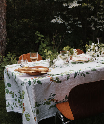 A table set outdoors with a Tomate Verte Linen Tablecloth, glassware, plates, and vases of fresh flowers, surrounded by wooden chairs and lush greenery.