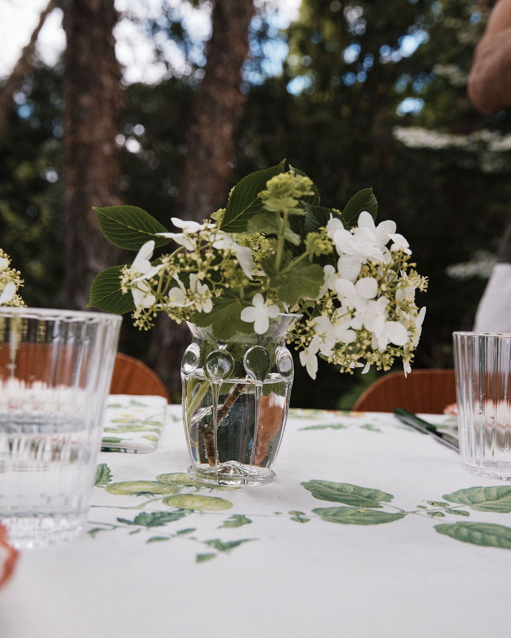 A small, handcrafted Carlotta Vase filled with white flowers sits on a table covered by a floral tablecloth. Two clear drinking glasses are nearby, with trees visible in the blurred background.
