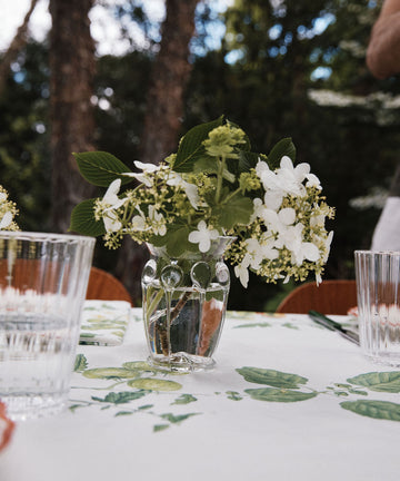 A small, handcrafted Carlotta Vase filled with white flowers sits on a table covered by a floral tablecloth. Two clear drinking glasses are nearby, with trees visible in the blurred background.